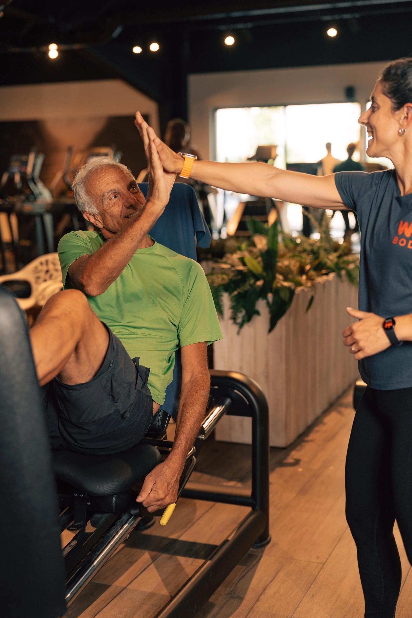 Un senior en tee-shirt vert s'entraîne sur un vélo de fitness en salle de fitness à Saint-Orens avec un coach