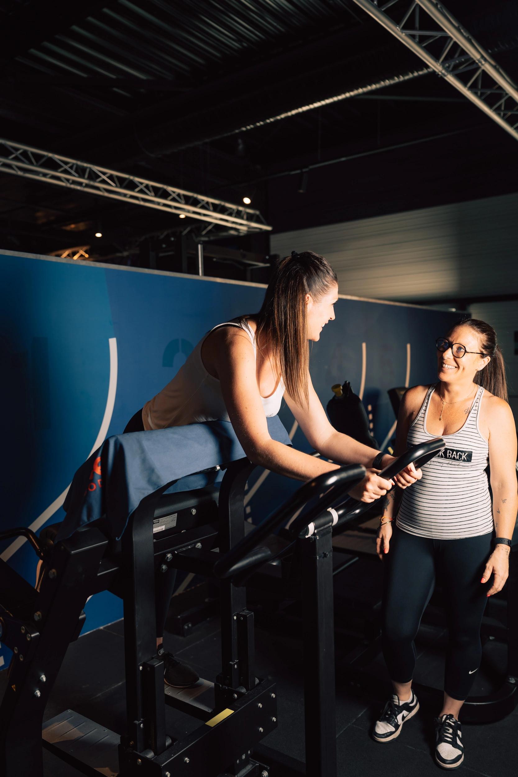 Une femme coach assiste une sportive sur un vélo stationnaire en salle de sport à Saint-Orens